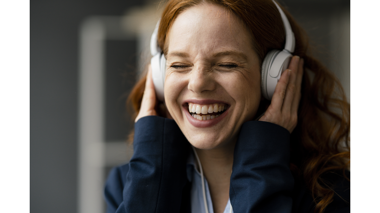 Portrait of laughing redheaded businesswoman listening music with white headphones