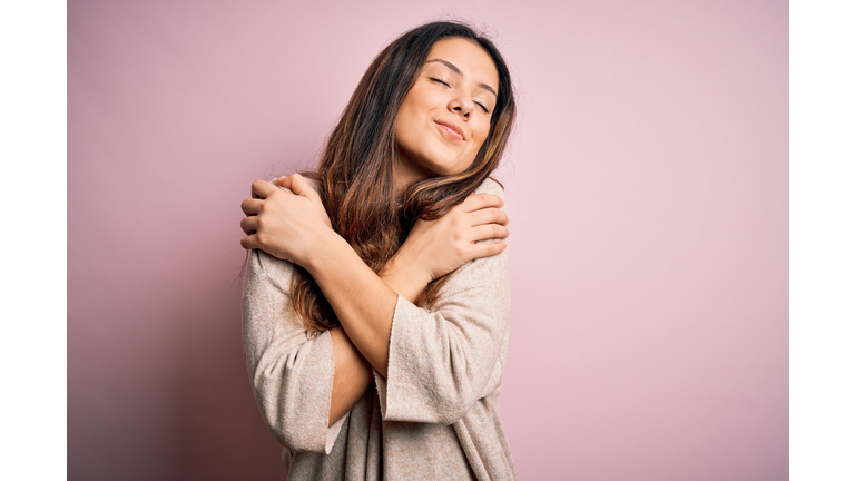 Young beautiful brunette woman wearing casual sweater standing over pink background Hugging oneself happy and positive, smiling confident. Self love and self care