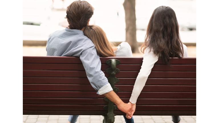 Boyfriend Holding Hands With Girlfriend's Friend Sitting On Bench Outdoor