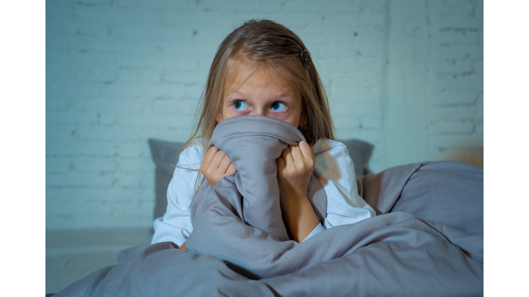 Scared little girl staying sleepless hiding behind the duvet looking horrified in the dark having childhood nightmares in child imagination Sleeping disorders Stress Depression and Insomnia concept.