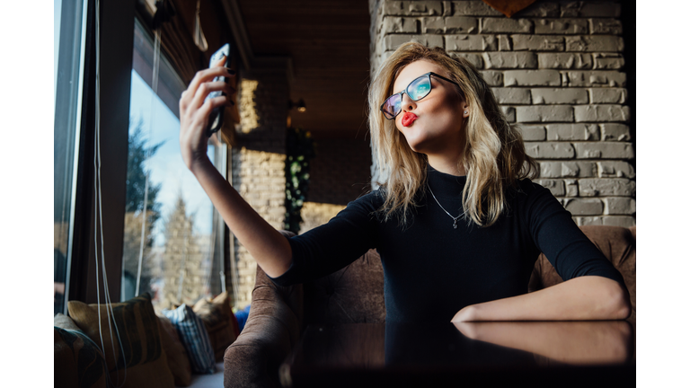 Young beautiful blonde woman taking a selfie in coffee shop. Hipster, red lips, glasses