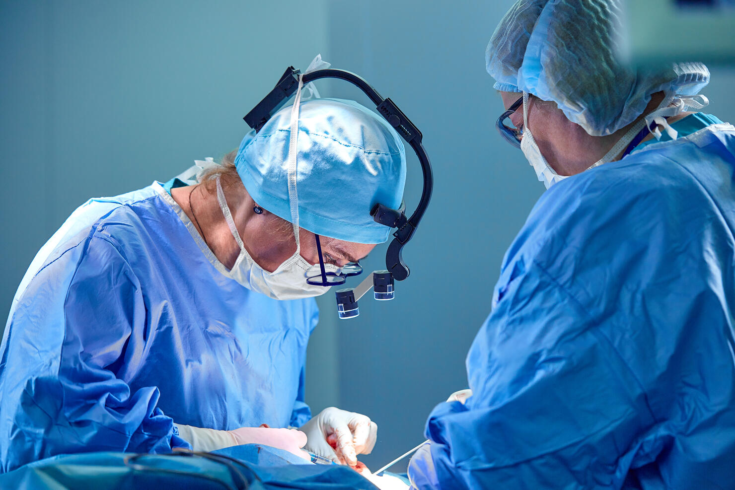 Surgeon and his assistant performing cosmetic surgery in hospital operating room. Surgeon in mask wearing loupes during medical procadure