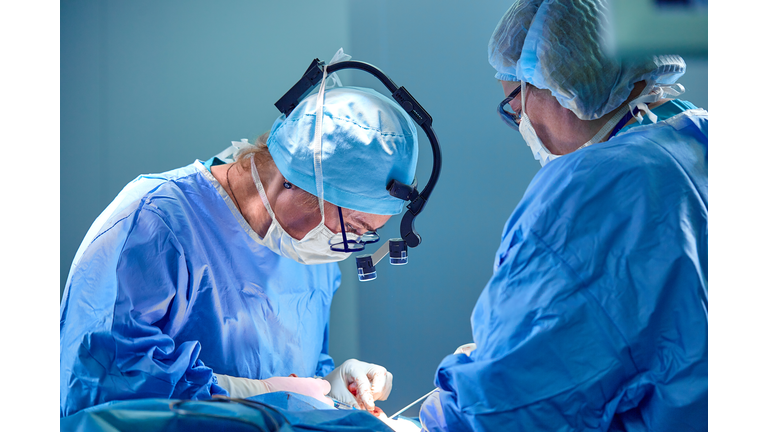 Surgeon and his assistant performing cosmetic surgery in hospital operating room. Surgeon in mask wearing loupes during medical procadure