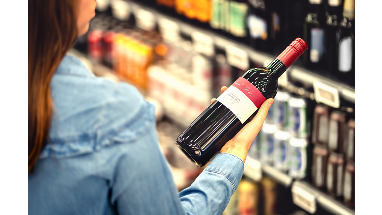 Woman reading the label of red wine bottle in liquor store or alcohol section of supermarket. Shelf full of alcoholic beverages. Female customer holding and choosing a bottle of merlot.