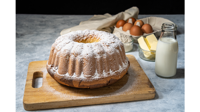 Fresh bundt cake decorated with icing sugar on a wooden cutting board with ingredients in a background. Front view of a delicious homemade marble cake baked in a mold.