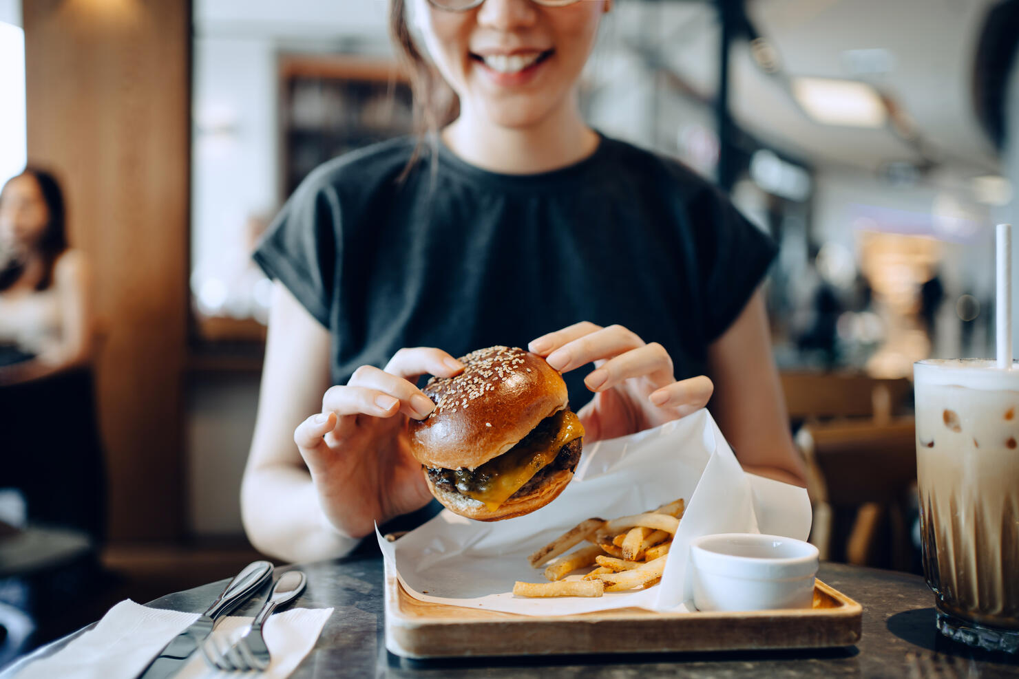 Smiling young Asian woman enjoying freshly made delicious burger with fries and a glass of iced coffee in a cafe