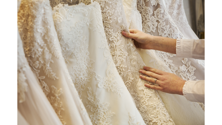 Rows of wedding dresses on display in a specialist wedding dress shop. Close up of full skirts, some with a lace overlay, in a variety of colour tones. .