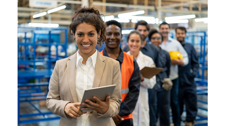 Beautiful female manager at a factory holding a tablet and team of blue collar workers, engineers and inspectors standing in a row smiling at camera