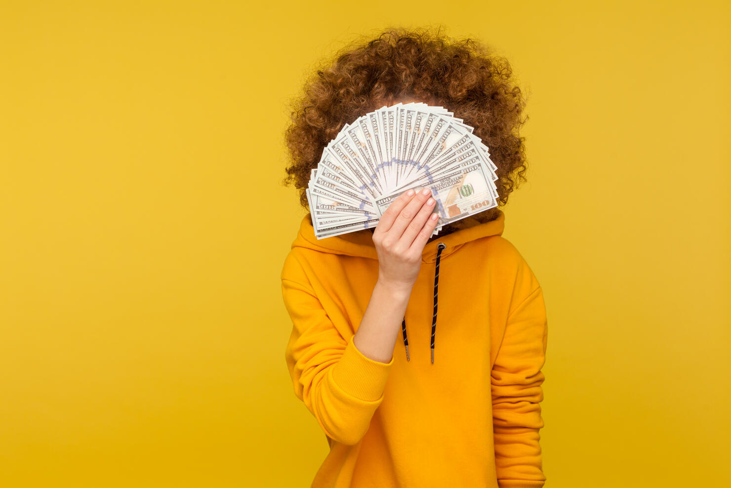 Curly-haired woman in urban style hoodie hiding face behind bunch of dollar banknotes, anonymous person