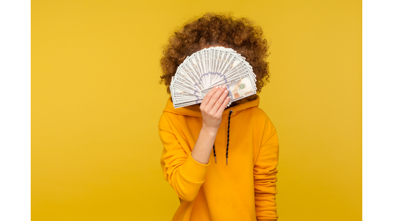 Curly-haired woman in urban style hoodie hiding face behind bunch of dollar banknotes, anonymous person