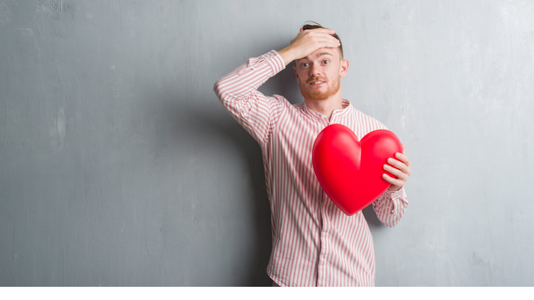 Young redhead man over grey grunge wall holding red heart stressed with hand on head, shocked with shame and surprise face, angry and frustrated. Fear and upset for mistake.