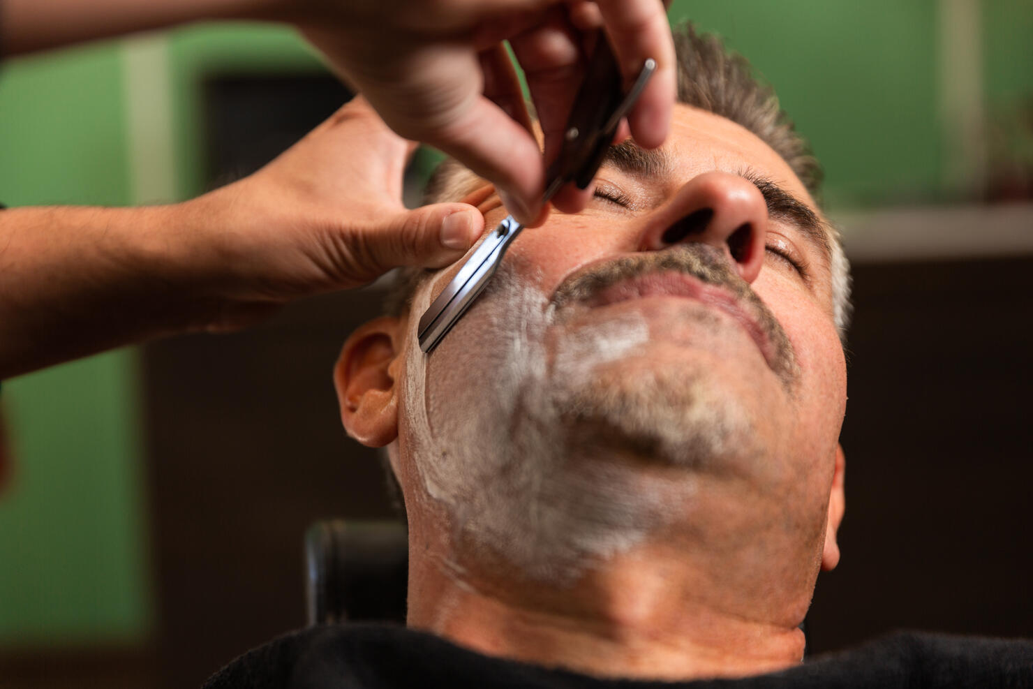 barber shaves a customer's cheek full of shaving foam with a razor