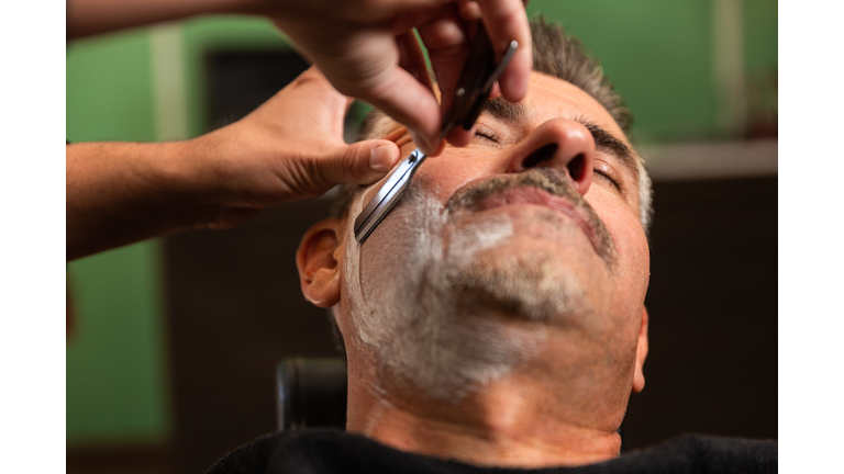 barber shaves a customer's cheek full of shaving foam with a razor