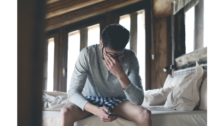 Tired young man with smartphone sitting on bed at home squinting