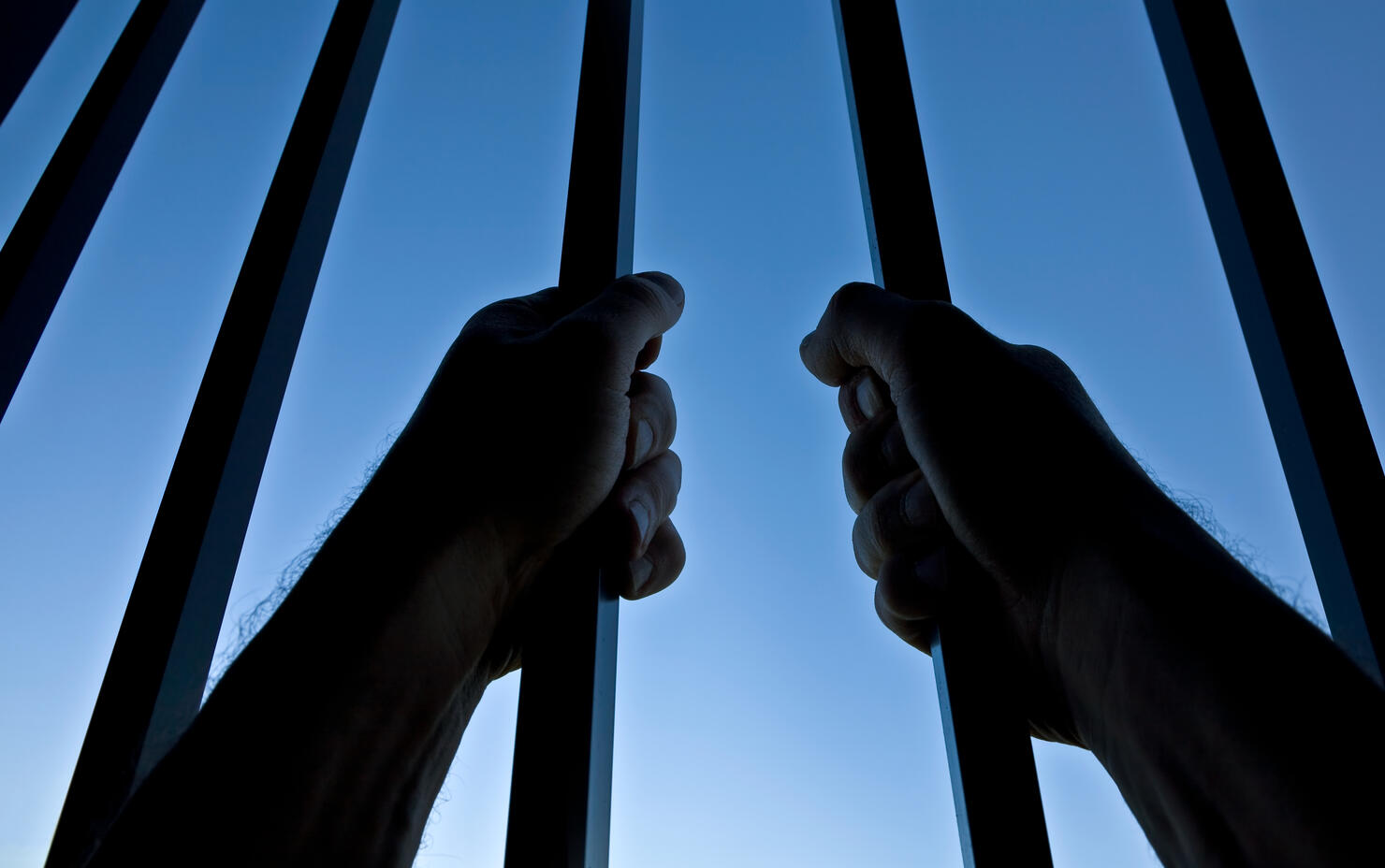 Silhouette of Hands Behind Jail Bars Against Clear Blue Sky
