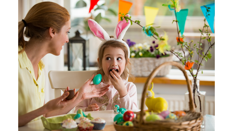 Mother and daughter celebrating Easter, eating chocolate eggs. Happy family holiday. Cute little girl in bunny ears laughing, smiling and having fun.