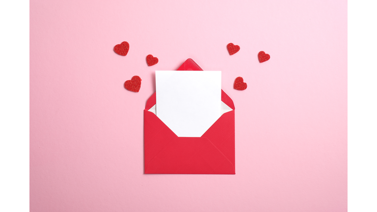 Red paper envelope with blank white note mockup inside and Valentines hearts on pink background. Flat lay, top view. Romantic love letter for Valentine's day concept.