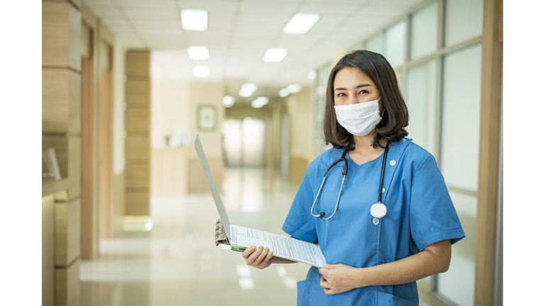 Shot of Asian Female nurse holding a medical record in an hospital corridor. Her wearing protective face mask and hospital gown for prevention infection virus.