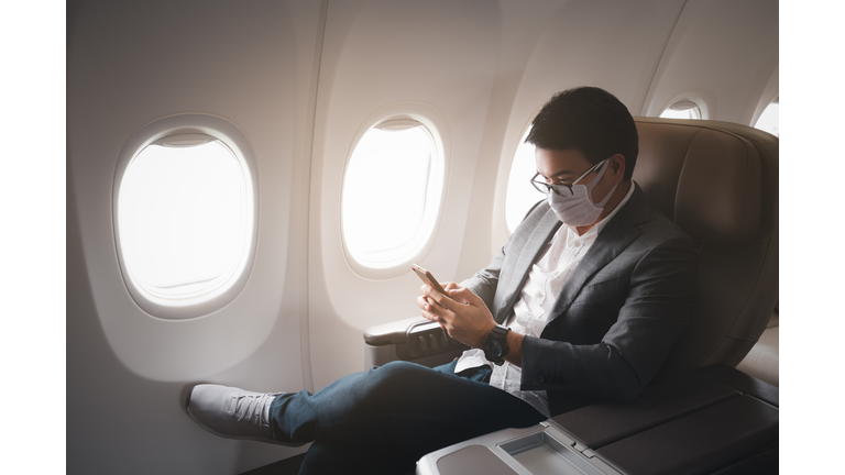 Young asian businessman wearing protective face mask with suit sitting in business first class seat using smartphone due to Coronavirus or COVID-19 outbreak situation in all of landmass in the world