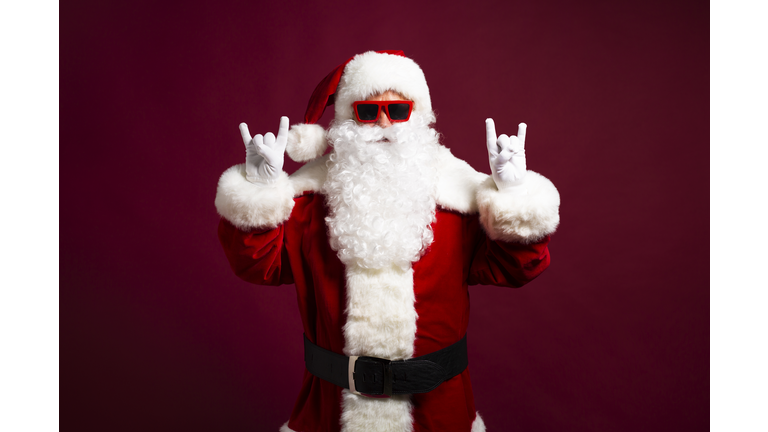 Stylish man in Santa Clause costume and sunglasses looking at camera and showing rock sign gesture