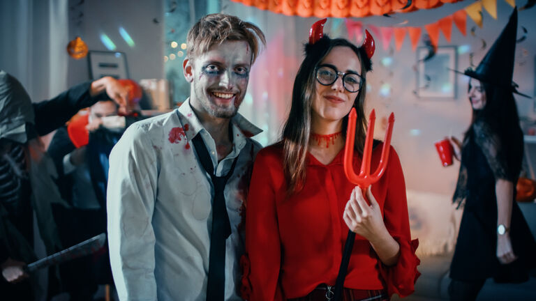 Halloween Costume Party: Brain Dead Zombie and Beautiful She Devil with Trident Pose as a Couple. In the Background Monsters Having Fun and Dancing in the Decorated Room