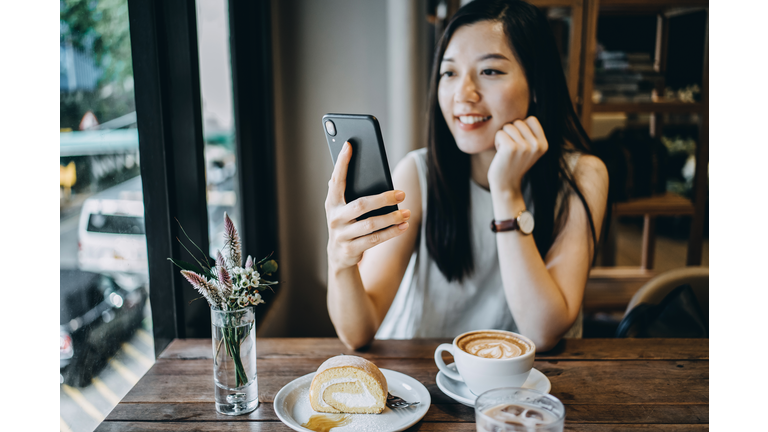 Smiling young woman using smartphone while having a relaxing time enjoying coffee and cake in a coffee shop