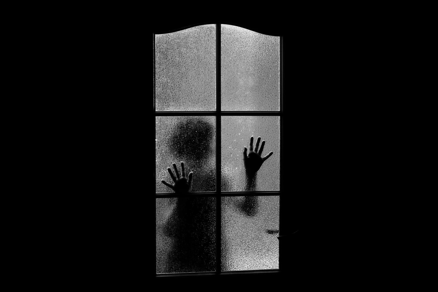 Dark silhouette of girl behind glass. Locked alone in room behind door on Halloween in grayscale. Nightmare of child with aliens, monsters and ghosts. Evil in home in monochrome. Inside haunted house.