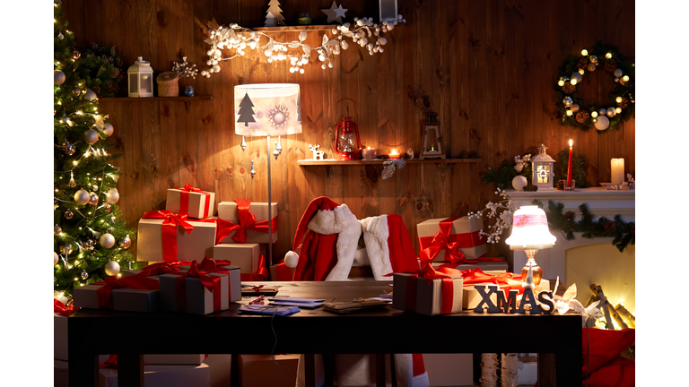 Santa Claus costume and hat hanging on chair at table with Merry Christmas decor gifts presents on holiday eve in cozy Santa home workshop interior late in night with light on xmas tree and fireplace.