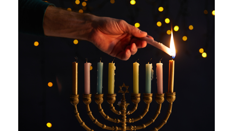 partial view of man lighting up candles in menorah on black background with bokeh lights on Hanukkah