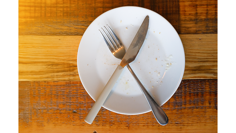 Cutlery On A Dirty Used White Plate, On A Wooden Background. The End Of The Lunch, Dinner Or Breakfast. Fork And Spoon In A Bowl. The Concept Of Food. Copy The Space For The Text.