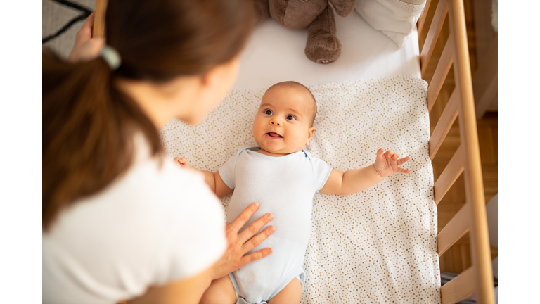 Mother putting her baby to sleep. Woman bending forward over a crib to check her baby.