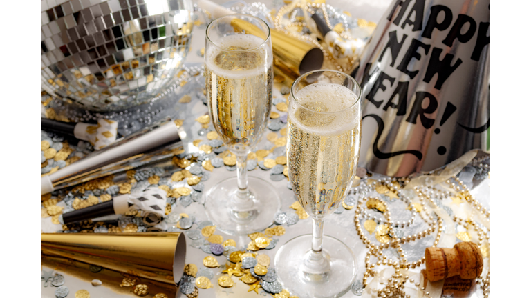 Happy new year celebration concept theme with close up on two glasses of champagne, disco ball covered in mirror, noise makers and party trumpets, confetti, beads and a cork on silver background