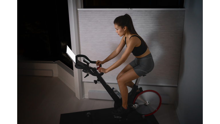 Home workout indoor stationary bike Asian girl biking screen with online classes woman training on smart fitness equipment indoors for cycling exercise. Late at night in bedroom