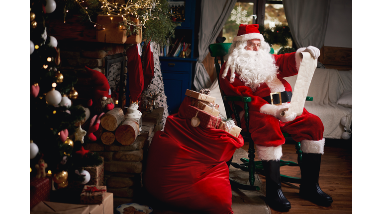 Portrait of Santa Claus, sitting in chair with sack full of presents, looking at Christmas list