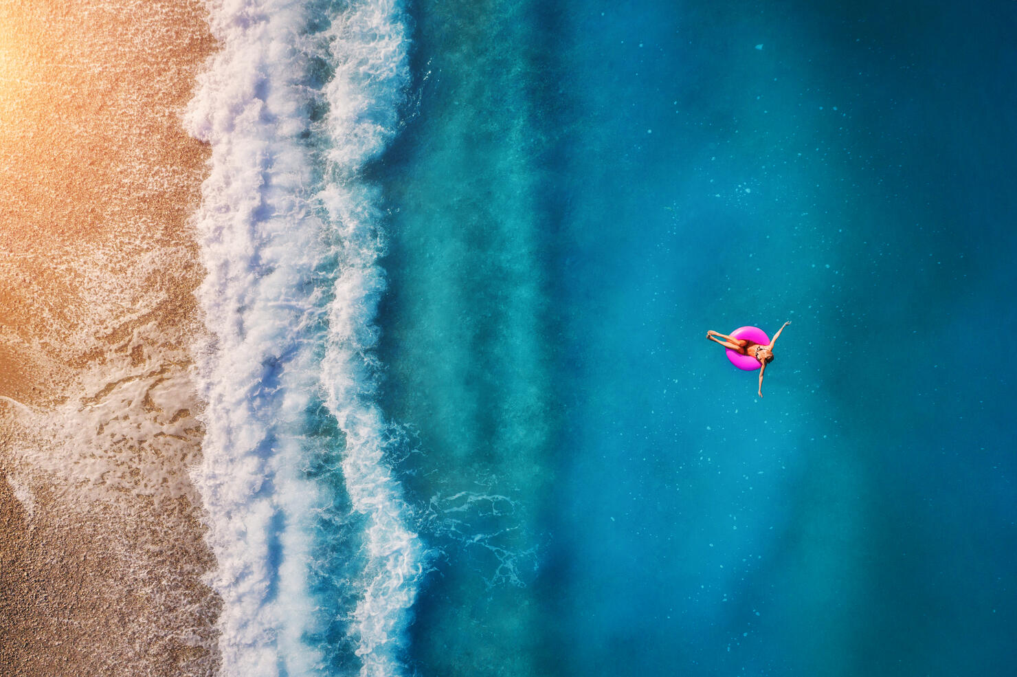 Aerial view of young woman swimming on the pink swim ring in the transparent turquoise sea in Oludeniz. Summer seascape with girl, beach, beautiful waves, blue water at sunset. Top view from drone