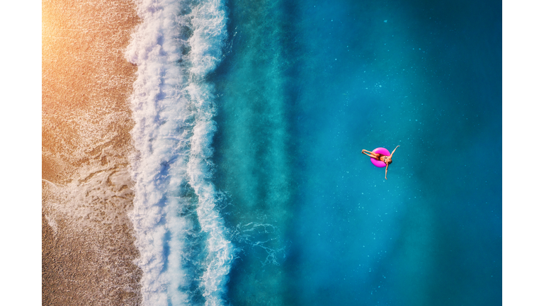 Aerial view of young woman swimming on the pink swim ring in the transparent turquoise sea in Oludeniz. Summer seascape with girl, beach, beautiful waves, blue water at sunset. Top view from drone