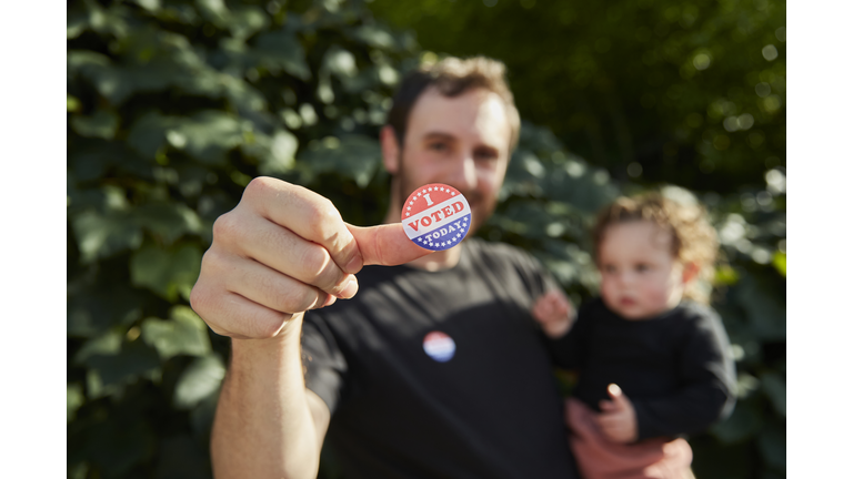 A father standing outside holding his baby, with an "I Voted Sticker" on his thumb