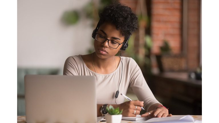 Focused mixed race woman wearing headphones watching webinar write notes