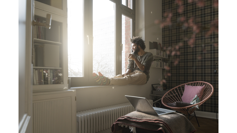 Man sitting on window sill in living room looking outside holding a cup