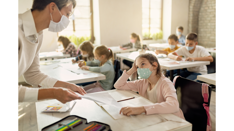 Schoolgirl with face mask talking to a teacher while receiving test results in the classroom.