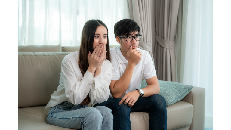 Asian Couple man and woman watching and enjoying terror tv movie sitting on a couch together in the livingroom at home. Family lifestyle relax and recreation concept.