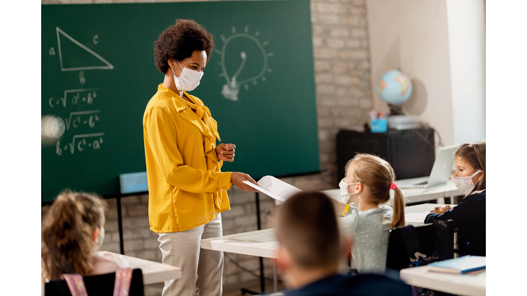 Happy black teacher giving exam paper to her student while wearing protective face mask in the classroom.