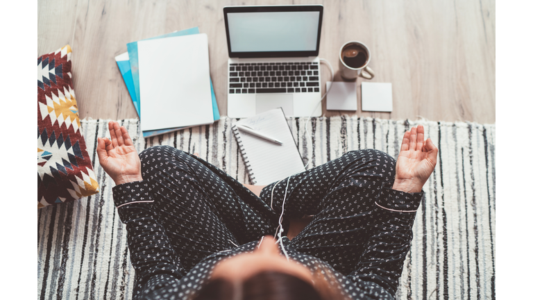 Businesswoman dressed in pajamas, listening earphones meditating with morning coffee with Gyan Mudra palm on floor office with laptop, papers top view shot. Distance work in quarantine time concept.