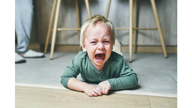 A little boy with blond hair, two years old, is lying on the floor and crying hysterically after kindergarten at home. Parenting.