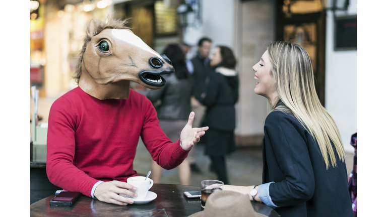 Woman and horse face man with costume drinking coffee in bar terrace and talking having fun