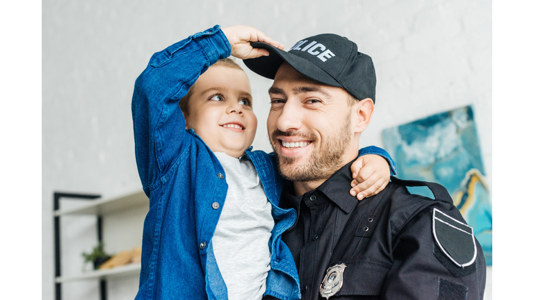 close-up portrait of smiling young father in police uniform carrying his little son and looking at camera