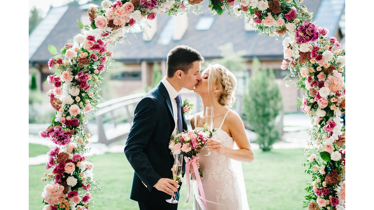 The bride and groom kissing. Newlyweds with a wedding bouquet, holding glasses of champagne standing on wedding ceremony under the arch decorated with flowers and greenery of the outdoor.