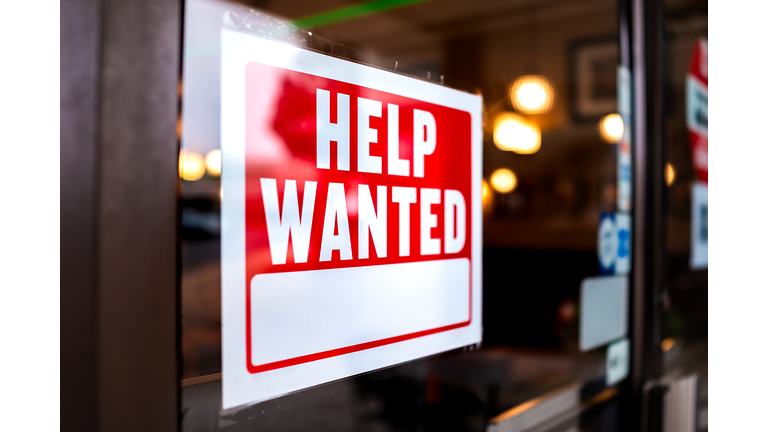 Sign text closeup for help wanted with red and white colors by entrance to store shop business building during corona virus covid 19 pandemic