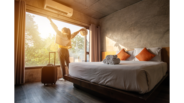 Portrait of tourist woman raised her hands and standing nearly window, looking to beautiful view with her luggage in hotel bedroom after check-in.