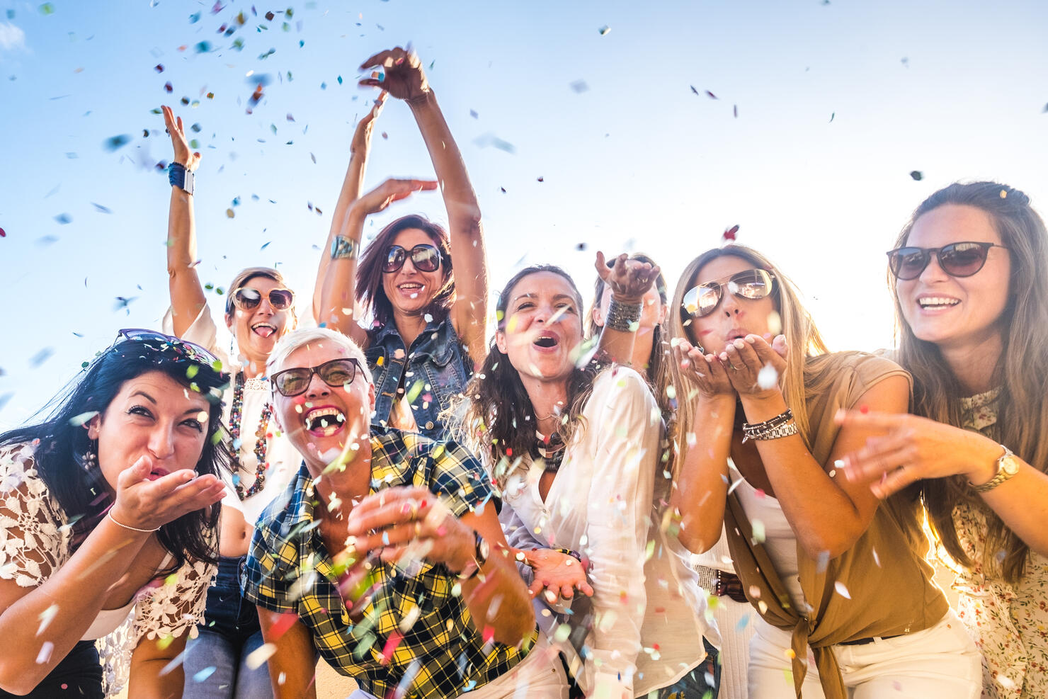 Cheerful group of happy people mixed ages generations women having fun all together during celebration party or carnival - view of fiends blowing. coloured confetti and laugh a lot in friendship